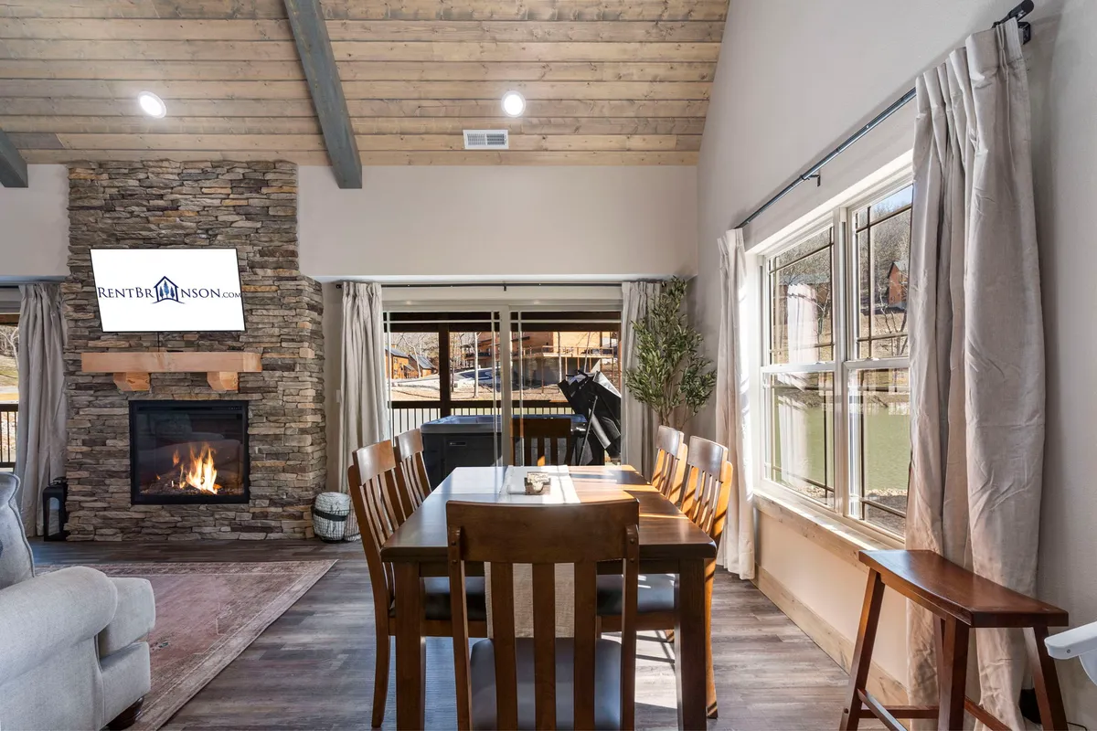 A welcoming interior view with a stone fireplace, dining table set for family meals, large windows letting in natural light, and a television mounted on the wall. The layout highlights the seamless connection between dining, entertainment, and relaxation areas.