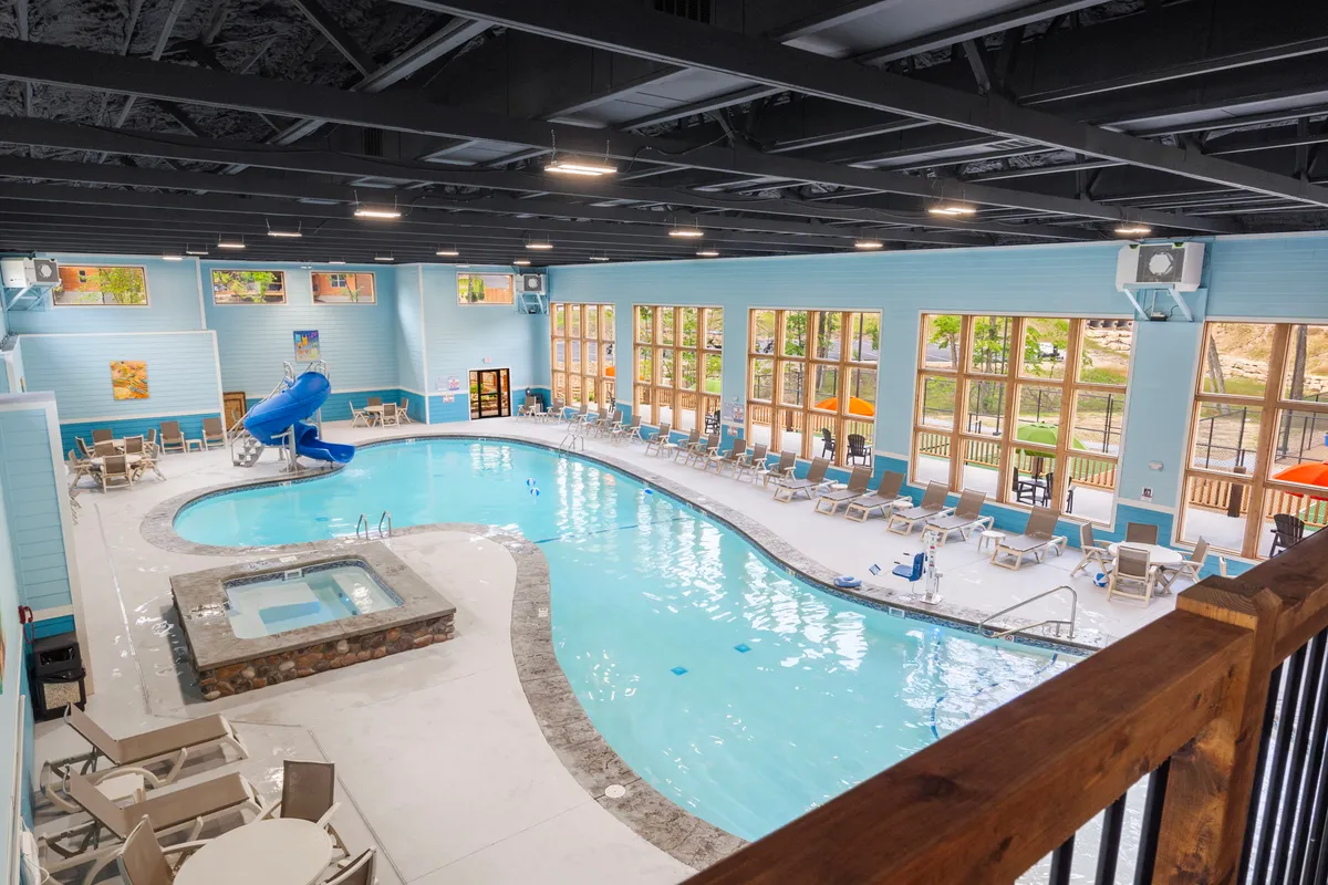 A vibrant indoor pool area featuring a large swimming pool, a fun water slide, a hot tub, and rows of poolside lounge chairs. Large windows let in natural light, and the space is set up for both swimming and relaxation, ideal for families and groups.