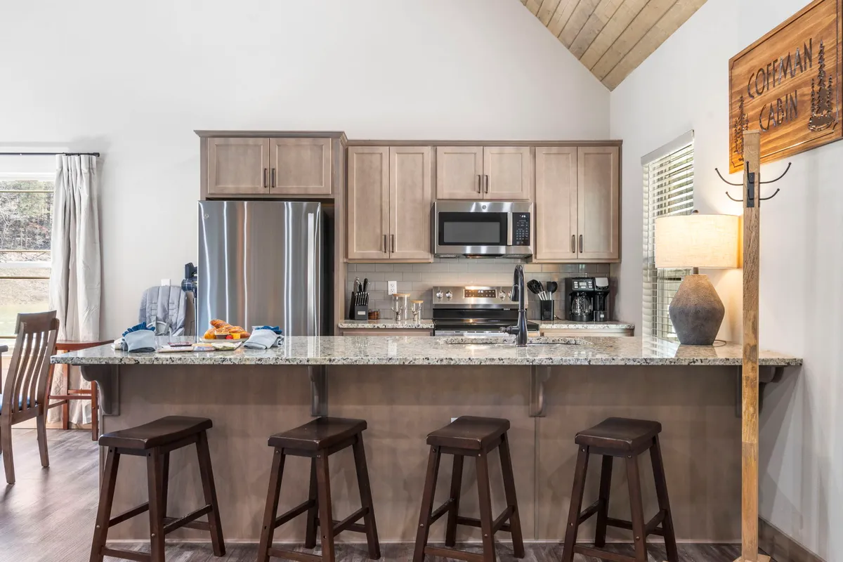 A modern chef-ready kitchen with granite countertops, stainless steel appliances, wooden bar stools at the island, and a decorative lamp. The kitchen is clearly integrated with the surrounding living and dining space, ideal for group meal prep and socializing.
