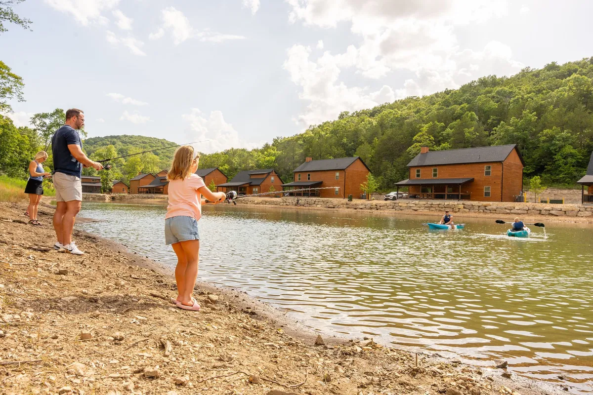 Families and children are seen fishing and playing by a scenic lake, with cozy cabins in the background. The mood is joyful and communal, with lots of outdoor activity and natural beauty.