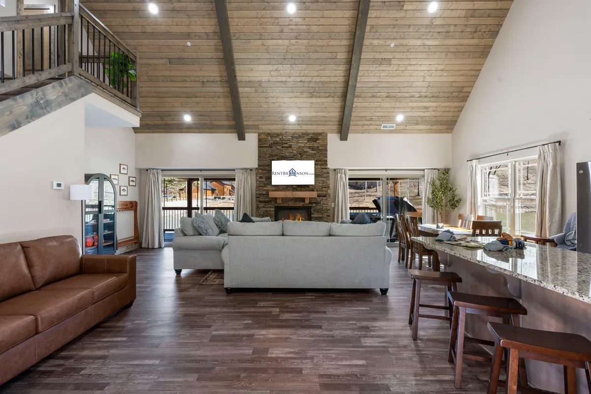A spacious open-concept living area featuring a plush sofa arranged around a stone fireplace. The adjoining dining area is visible, with large windows offering views of the Ozarks’ natural beauty. The space exudes warmth and invites social gatherings and relaxation.