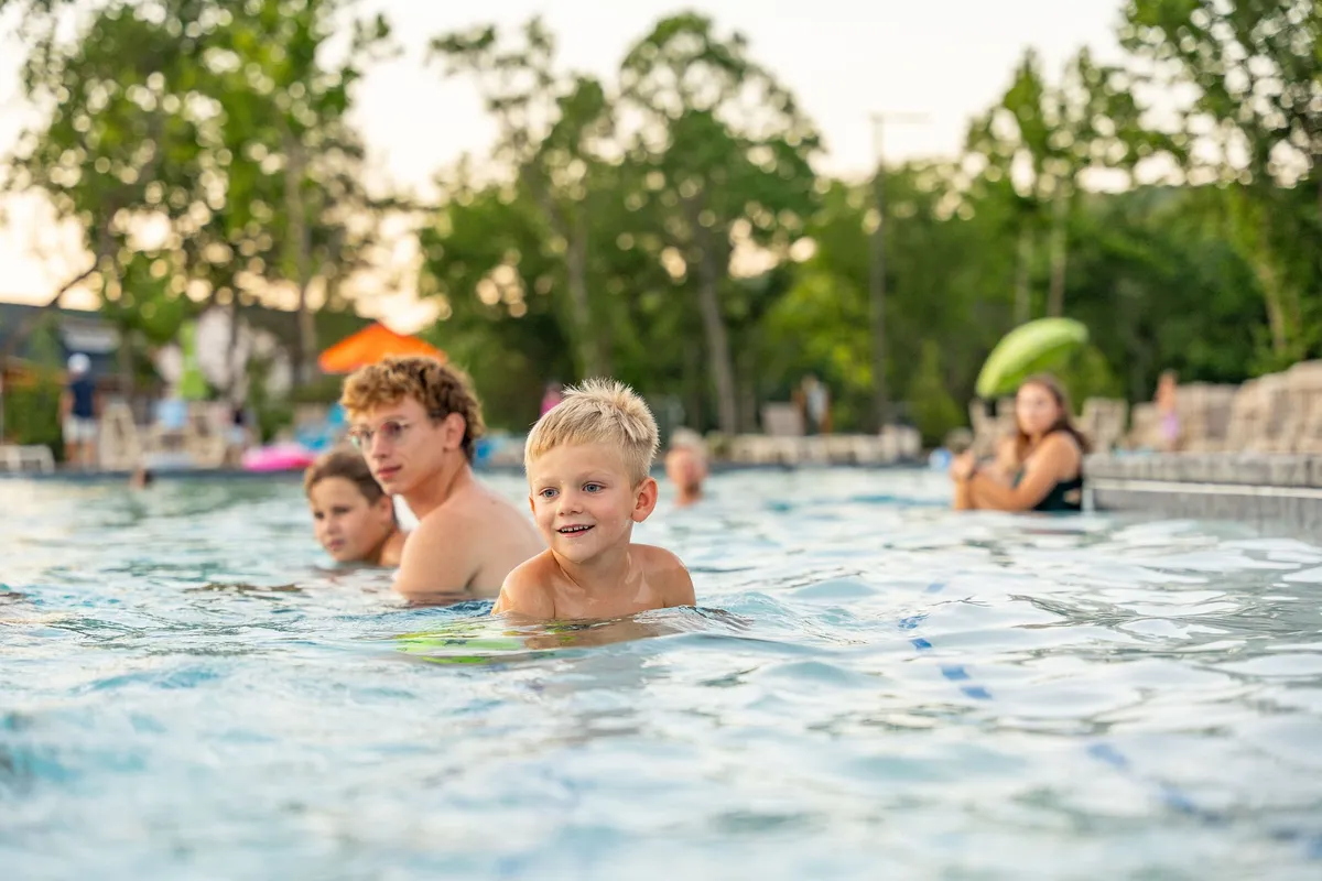 Children splash and swim in a pool while adults lounge nearby, enjoying the sun. The pool deck is lively and playful, with pool toys and carefree smiles creating a classic summer family scene.