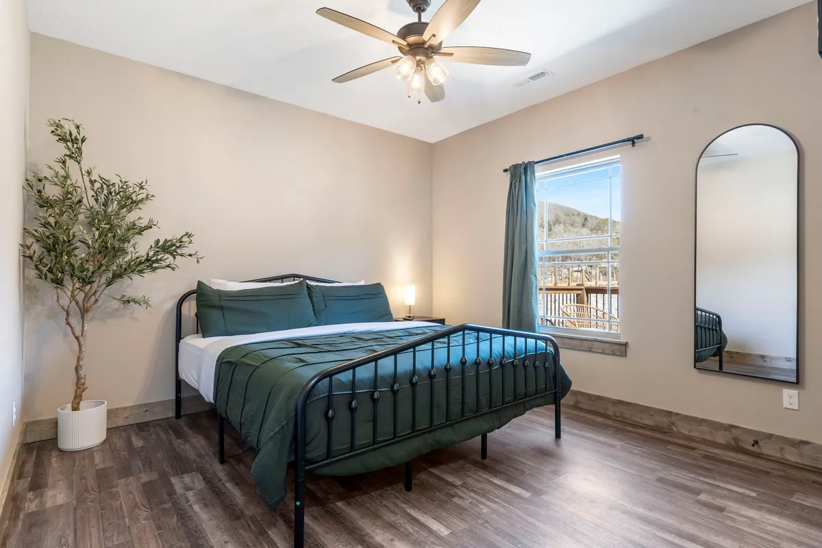 A serene bedroom featuring a plush bed with dark green bedding, a large mirror reflecting soft light, and a potted plant that adds a touch of nature. The space is uncluttered and calming, with natural light streaming in, ideal for relaxation and privacy.