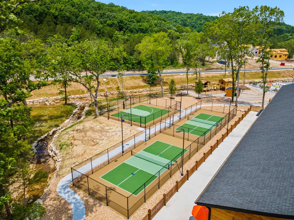 Active tennis and pickleball courts are surrounded by trees, with a playground and a walking path nearby. The space is alive with recreational possibilities, welcoming families to enjoy games and the outdoors together.
