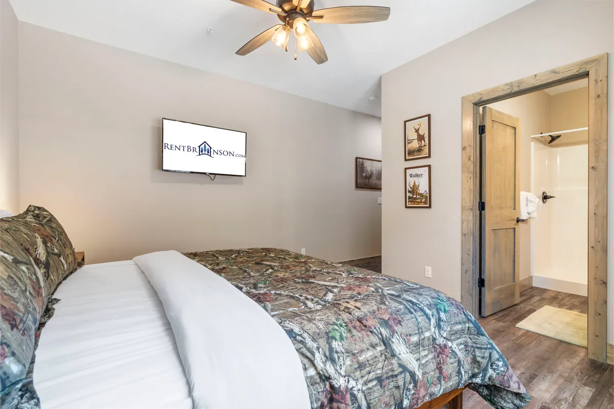 A rustic yet comfortable bedroom, featuring camouflage bedding, a wall-mounted TV, decorative wall art, and a bathroom door. The design exudes warmth and individuality, emphasizing privacy and relaxation.