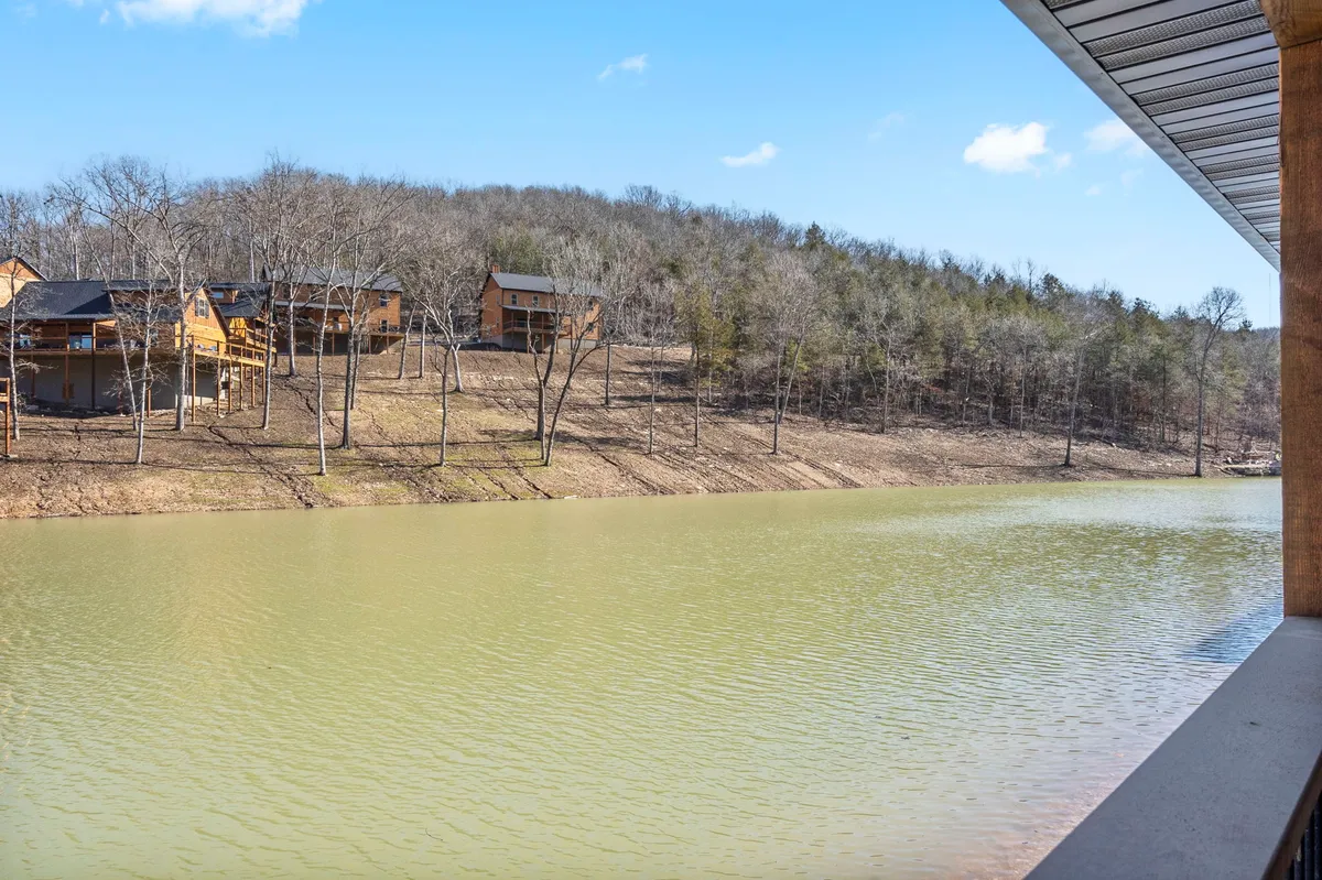 A wide, tranquil view of a calm lake bordered by wooden cabins and a tree-covered hillside. The setting radiates peace and offers opportunities for outdoor activities like picnicking, wildlife watching, and lakeside relaxation.